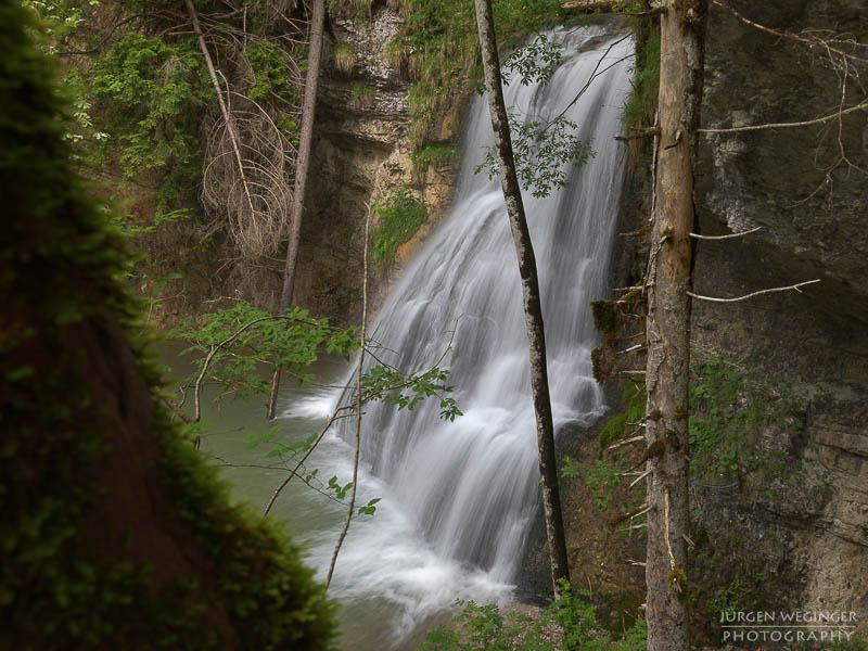 Wasserfall in einem dichten Wald mit moosbedeckten Felsen und grünem Laub