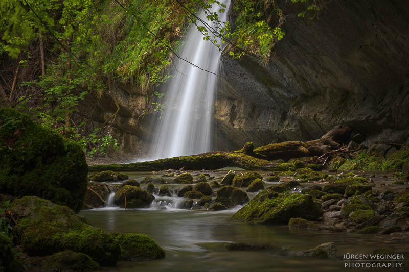 Wasserfall in einem dichten Wald mit moosbedeckten Felsen und grünem Laub