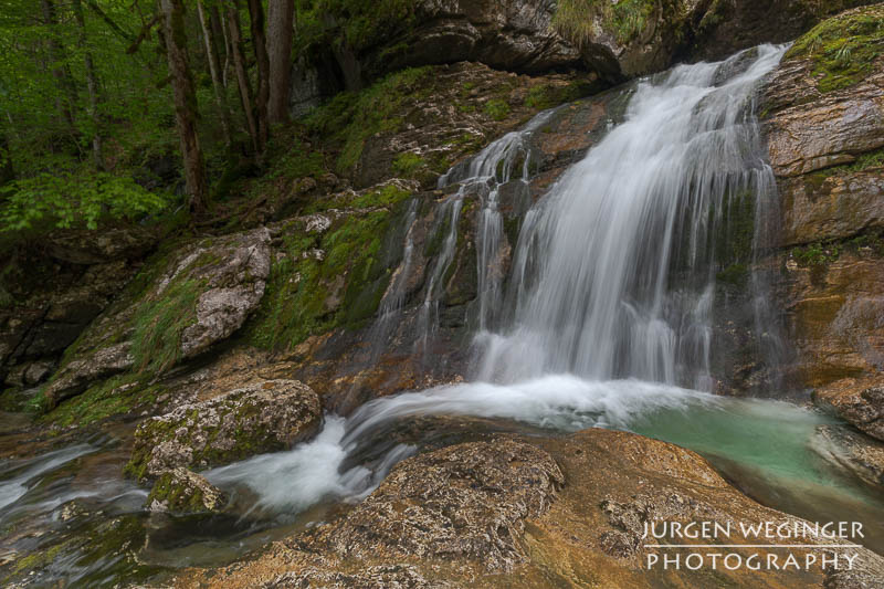 Ein Wasserfall fließt über Felsen, der von einer grünen bewaldeten Landschaft umgeben ist.