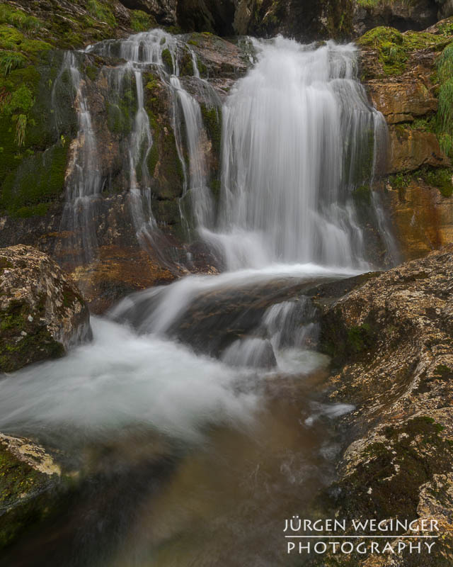 Ein Wasserfall fließt über Felsen, der von einer grünen bewaldeten Landschaft umgeben ist.