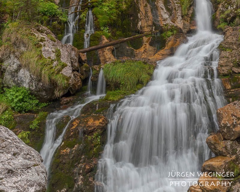 Ein Wasserfall fließt über Felsen, der von einer grünen bewaldeten Landschaft umgeben ist.