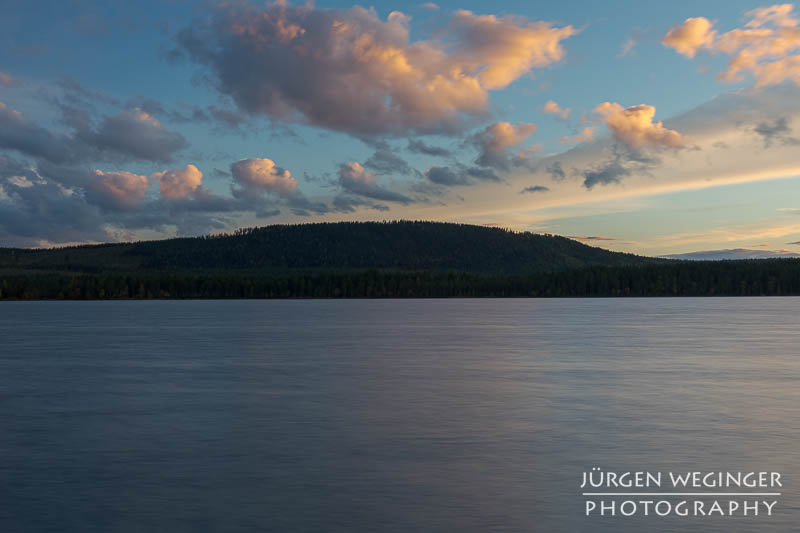 Ein See mit einem bewaldetem Hügel im Hintergrund. Blauer Himmel mit Wolken zu Sonnenuntergang