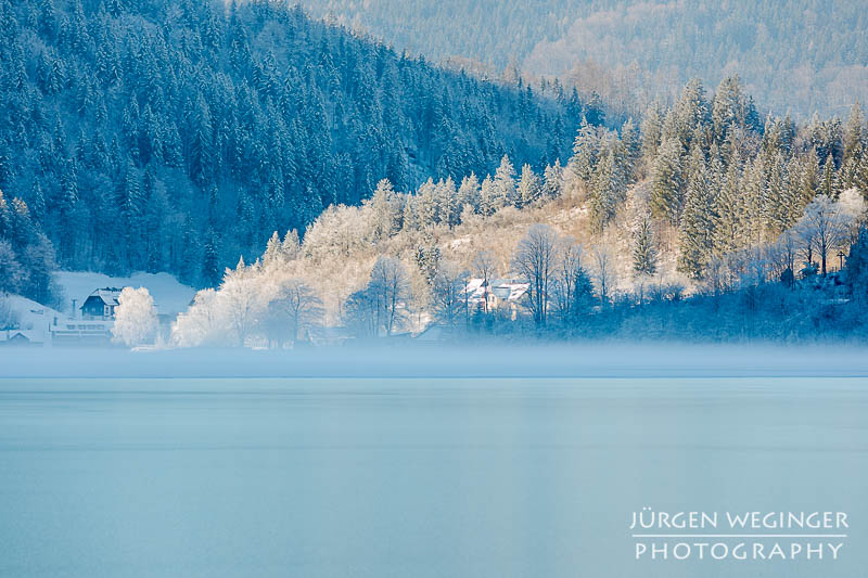 Nebel über einem See mit einem Sonnen beschienenen Waldstück im Hintergrund